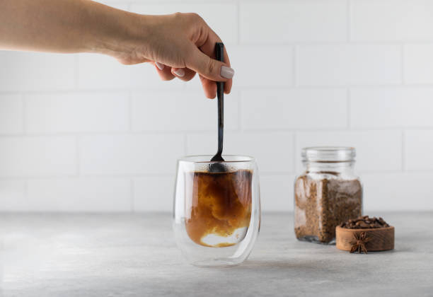 Woman stirs instant coffee in glass mug with boiled water on grey stone table Woman stirs instant granulated coffee in glass mug with hot boiled water by spoon on grey stone table in light kitchen closeup glass cup of boiling water stock pictures, royalty-free photos & images