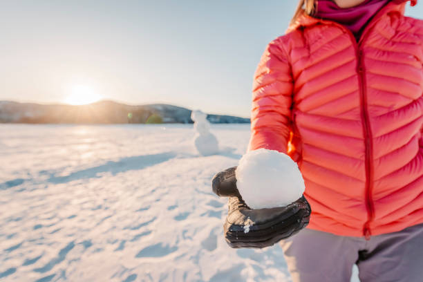 conceito de estilo de vida ativo ao ar livre de inverno. menina segurando bola de neve em dia de neve com boneco de neve no fundo em lago congelado no belo pôr do sol de inverno - snowball snow play throwing - fotografias e filmes do acervo