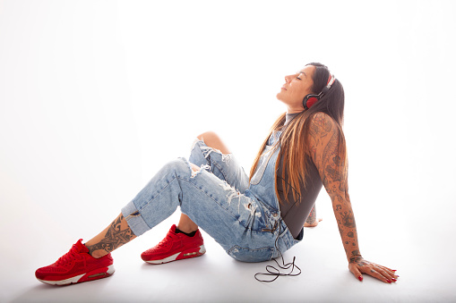 studio photography on white background of a tattooed young woman listening to music on her headphones, sitting in a relaxed attitude
