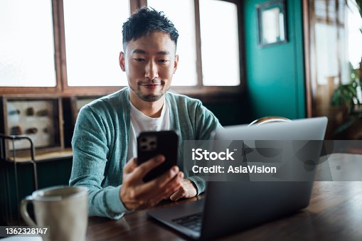 istock Confident young Asian man looking at smartphone while working on laptop computer in home office. Remote working, freelancer, small business concept 1362328147