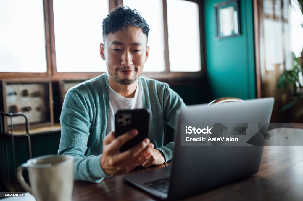 Confident young Asian man looking at smartphone while working on laptop computer in home office. Remote working, freelancer, small business concept Using Phone Stock Photo