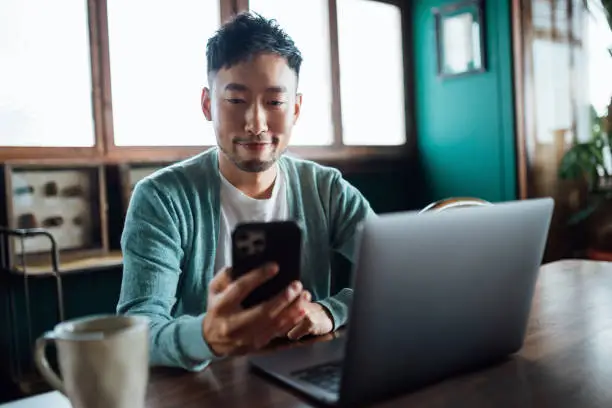 Photo of Confident young Asian man looking at smartphone while working on laptop computer in home office. Remote working, freelancer, small business concept