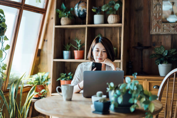 istock Busy young Asian woman working from home, using laptop computer while looking at smartphone sitting at desk at home office. Remote working, freelancer, small business concept 1362328149