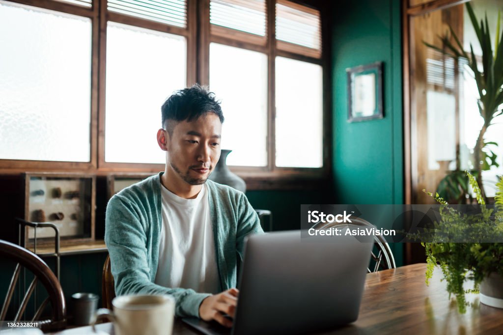 Professional young Asian man working from home, using laptop computer in home office. Remote working, freelancer, small business concept Working At Home Stock Photo
