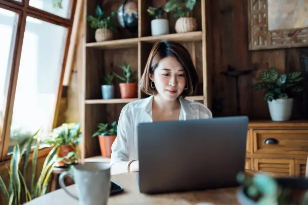 Young Asian woman working from home, video conferencing with business partners on laptop computer in home office. Remote working, freelancer, small business concept