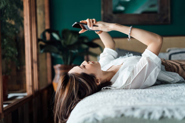 расслабленная молодая азиатская женщина проверяет социальные сети на смартфоне, лежа в постели. молодежная культура. технологии в повседн� - day in the life стоковые фото и изображения