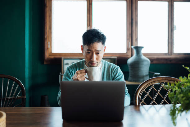 istock Professional young Asian businessman working from home, having a cup of coffee while using laptop computer in home office. Remote working, freelancer, small business concept 1362328345