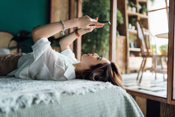 joven asiática relajada comprando en línea con un teléfono inteligente mientras está acostada en la cama. cultura juvenil. la tecnología en la vida cotidiana - lying down women laptop freedom fotografías e imágenes de stock