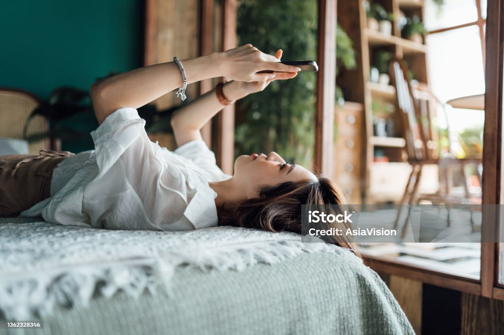 Relaxed young Asian woman shopping online with smartphone while lying in bed. Youth culture. Technology in everyday life Online Dating Stock Photo