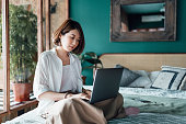 Young Asian woman working on laptop while sitting on bed in bedroom. Remote working, freelancer, small business concept