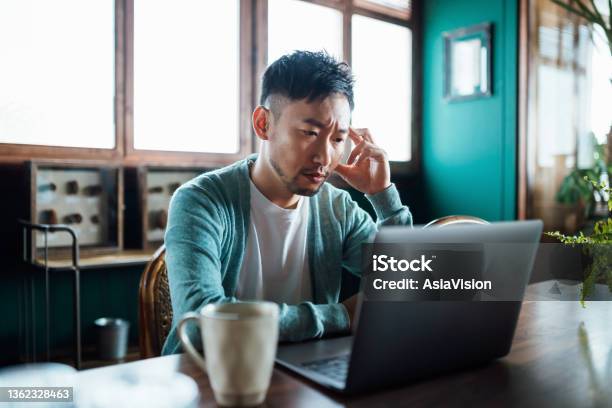 Worried Young Asian Man With His Hand On Head Using Laptop Computer At Home Looking Concerned And Stressed Out Stock Photo - Download Image Now