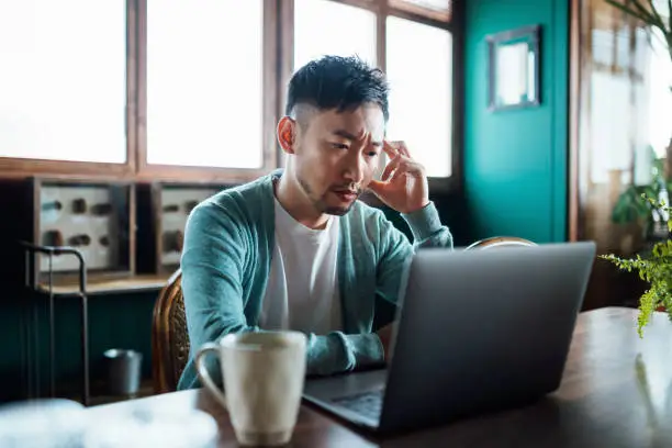 Photo of Worried young Asian man with his hand on head, using laptop computer at home, looking concerned and stressed out