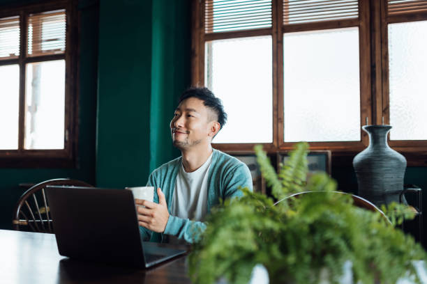 istock Contemplated young Asian man sitting at a table, using laptop computer with a cup of coffee on hand. He is in thought and looking away in home office. Remote working, freelancer, small business concept 1362328473
