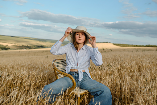 Young woman in casual outfit and a hat is sitting on a vintage chair in the wheat field. Freedom concept