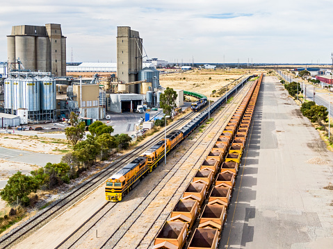 Port Adelaide, Australia - Dec 11, 2021: Aerial view busy railway yard with loaded iron ore train, empty ore wagons (freight cars) and approaching empty container train with green containers. Trains operated by One Rail Australia and Bowmans Rail.