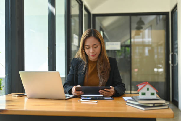 foto de uma jovem empresária segurando um tablet digital enquanto estava sentada na mesa de trabalho de madeira cercada por um laptop de computador, modelo de casa e vários equipamentos de escritório. - approved lease agreement insurance document - fotografias e filmes do acervo