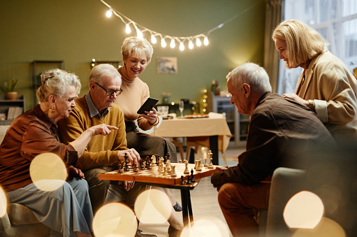 Three aged women watching two senior men playing chess during friends meeting in decorated living room