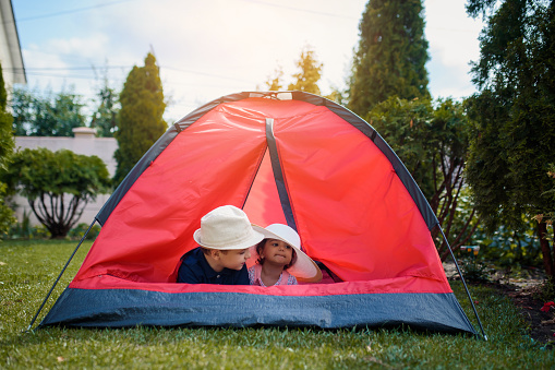 Two happy little kids boy and girl brother and sister are playing in a red camping tent in the home yard on the grass. Home camping vacation concept