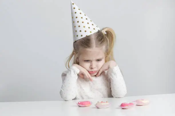 Photo of Displeased little girl with birthday hat and donuts in front of her.