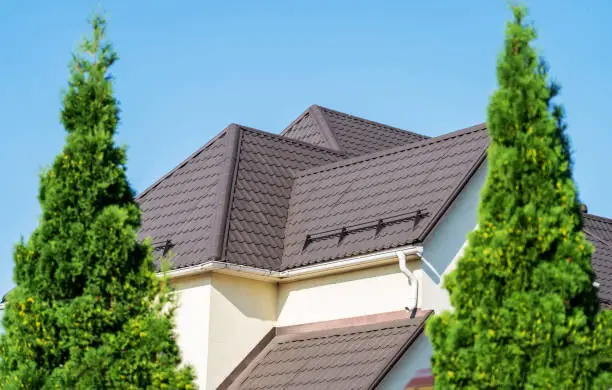Photo of Tiled roof with segment of snow holding structure, Gutter system for metal roof. House with new brown metal tile roof and rain gutter. Metallic Guttering System, Guttering and Drainage Pipe Exterior