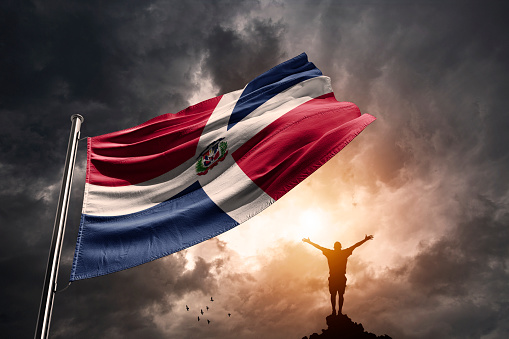 Dominican Republic flag in a sunset with dramatic storm clouds and a men raising his hands in victory.