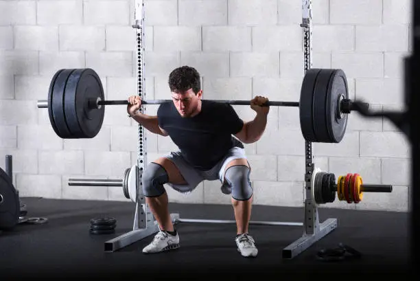 Full body view of young man performing back squats exercise in gym with heavy plates.