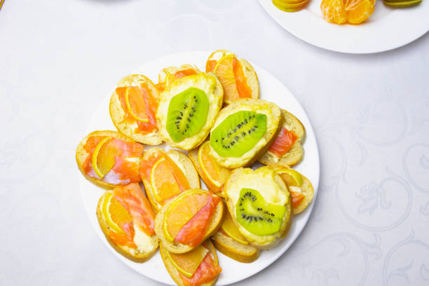 On the festive table, covered with a white tablecloth, there are sandwiches with red fish, cold cuts, fruits and vegetables