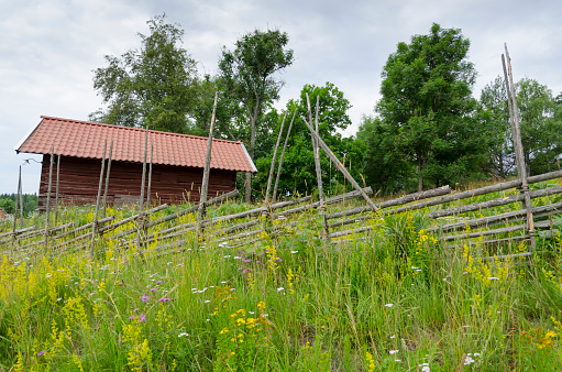 Historic old log cabin on an old farm in the Swedish countryside. The horizontal logs are interlocked at the corners by notching.
Typical Swedish round pole fence in the foreground.