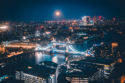 Panoramic aerial night and fullmoon view to the cityscape of London with Tower Bridge and the illuminated skyscrapers of the city during dusk in the United Kingdom