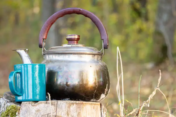 Photo of A cup of hot coffee near the burnt iron tea kettle on the tree stump. Adventure, travel, tourism and camping concept. Coffee cooked over a campfire on the nature. Selective focus