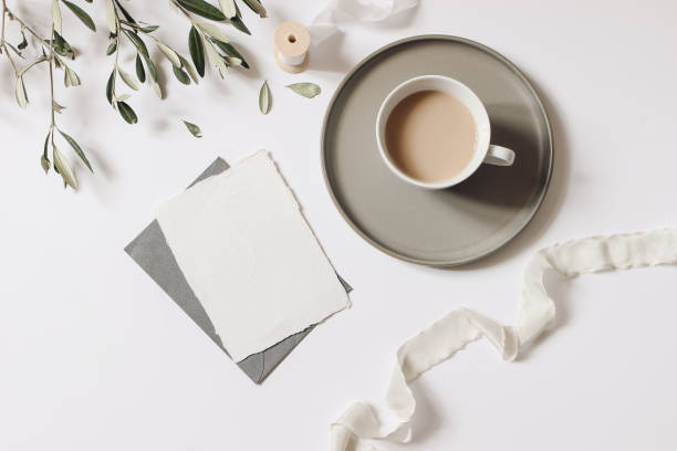 istock Summer still life. Cup of coffee, blank greeting card mockup with plate, envelope. Olive tree branch and silk ribbon, white table background. Wedding stationery. Flat lay, top view. Breakfast concept 1362421922