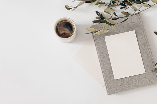 Summer still life. Cup of coffee, blank greeting card mockups with vintage book. Olive tree branch isolated on white table background. Mediterranean wedding stationery. Flat lay, top view, breakfast