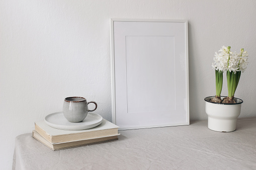 Spring breakfast still life scene. Cup of coffee, books. Empty white picture frame mockup. Beige linen tablecloth. Potted white hyacinth plant. Farmhouse, Scandinavian interior, Easter holiday concept.