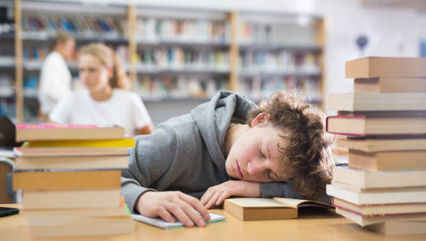 Tired teenager boy in library Tired teenager boy sleeping on table among books in library. boring homework twelve stock pictures, royalty-free photos & images