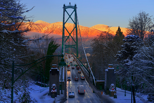 Top view of the bridge and moving cars with headlights on, orange mountains illuminated by the setting sun, blue sky, trees covered with snow