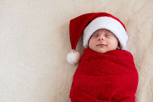 Top View Portrait First Days Life Newborn Cute Funny Sleeping Baby In Santa Hat Wrapped In Red Diaper At White Garland Background. Merry Christmas, Happy New Year, Infant, Childhood, Winter Copy space.