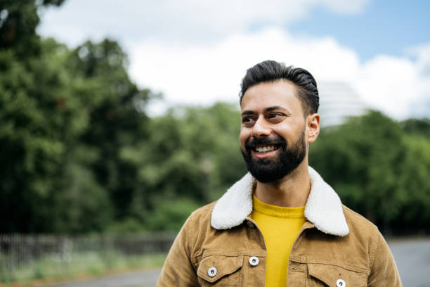 Candid portrait of early 30s Indian man standing outdoors Head and shoulders view of man with full beard in gold coloured sweater and tan jacket with shearing collar looking away from camera and smiling. candid stock pictures, royalty-free photos & images