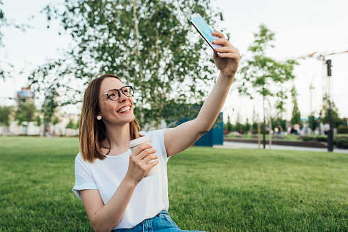 Portrait of a young woman relaxing in the park, enjoying coffee and taking a selfie.