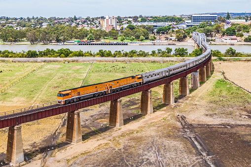 Aerial view luxury Overland Passenger Train crossing the Murray River en route to Melbourne, Sydney & Brisbane. Town business centre and houses on opposite riverbank, agricultural farmland and flood levee in foreground. ID & logos edited, paint on locomotive modified, graffiti removed from bridge.