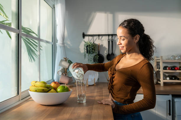 Young beautiful Latino woman pouring clean water into glass in kitchen. Attractive active thirsty girl drink or take a sips of mineral natural in cup for health care and wellbeing in kitchen in house. stock photo