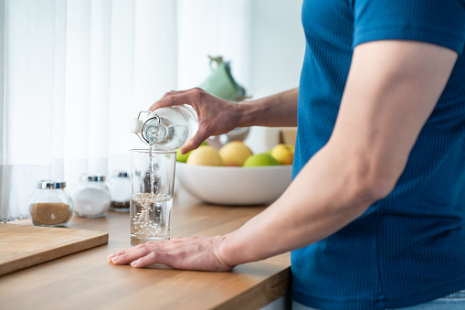 Close up hands of active strong man pouring clean water into glass. Attractive thirsty male drink or take a sip of mineral natural in cup after wake up for health and wellbeing in kitchen in house.
