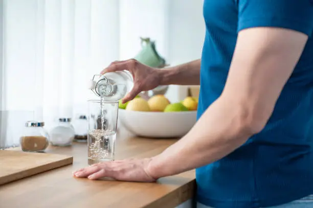 Photo of Close up hands of active strong man pouring clean water into glass. Attractive thirsty male drink or take a sip of mineral natural in cup after wake up for health and wellbeing in kitchen in house.
