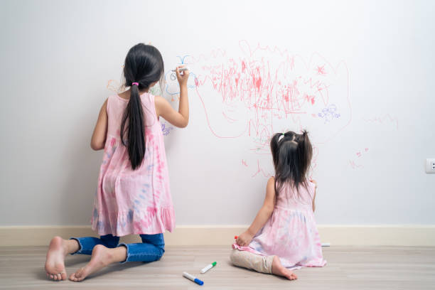 Asian young sibling kid girl enjoy paint on white wall in living room. Little adorable children having fun drawing and coloring art picture with hapiness enjoy creativity activity on holiday at home. stock photo