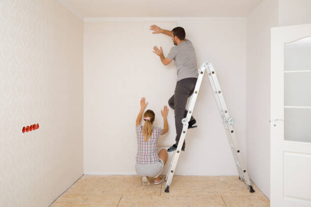 Young adult couple making interior change. Man standing on metal ladder and woman help applying new wallpaper on white wall in room. Working together. Repair work of home. Back view. stock photo