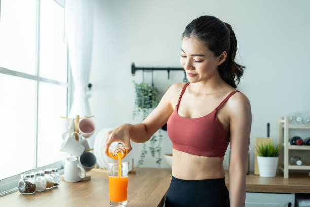 Portrait of Asian beautiful woman hold and pour orange juice in glass. Attractive sport girl in sportswear holding a cup of water, enjoy drink and eat healthy foods for health after exercise in house. stock photo