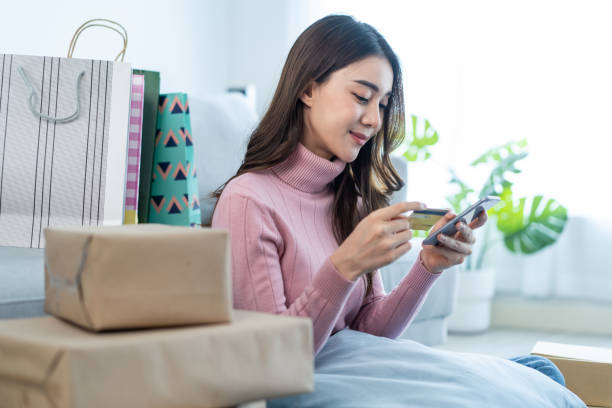 Asian attractive woman enjoy shopping online in living room at home. Beautiful girl sit on floor, hold credit card to make e-bank online payment after use laptop purchase goods in web store in house. stock photo