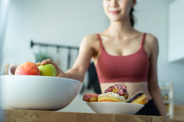 Asian attractive active woman pick up a green apple in kitchen at home. Beautiful sport girl in sportswear desire to chose fruit and ignore sweet donut enjoy eat healthy food for health after exercise stock photo