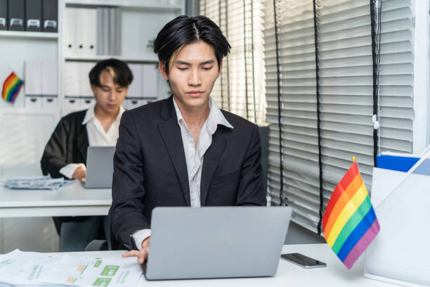 asian handsome businessman gay people working in office with happiness. attractive male lgbtq collegues in formal wear sitting on table, use laptop computer with gay pride and confidence at workplace. - homosexual gay pride business rainbow imagens e fotografias de stock