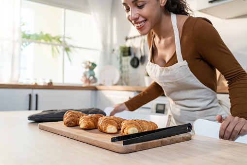 Attractive young Latino woman baking croissant on table in kitchen. Beautiful female wear apron feel happy and enjoy spending leisure time cooking bakery at home. Activity homemade in house concept.