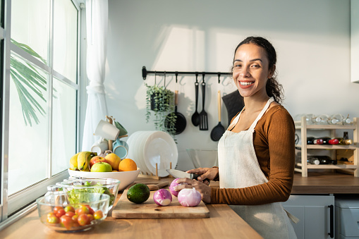 Latino attractive woman wear apron cook green salad in kitchen at home. Young beautiful girl feeling happy and enjoy eating vegetables healthy foods to diet and lose weight for health care in house.